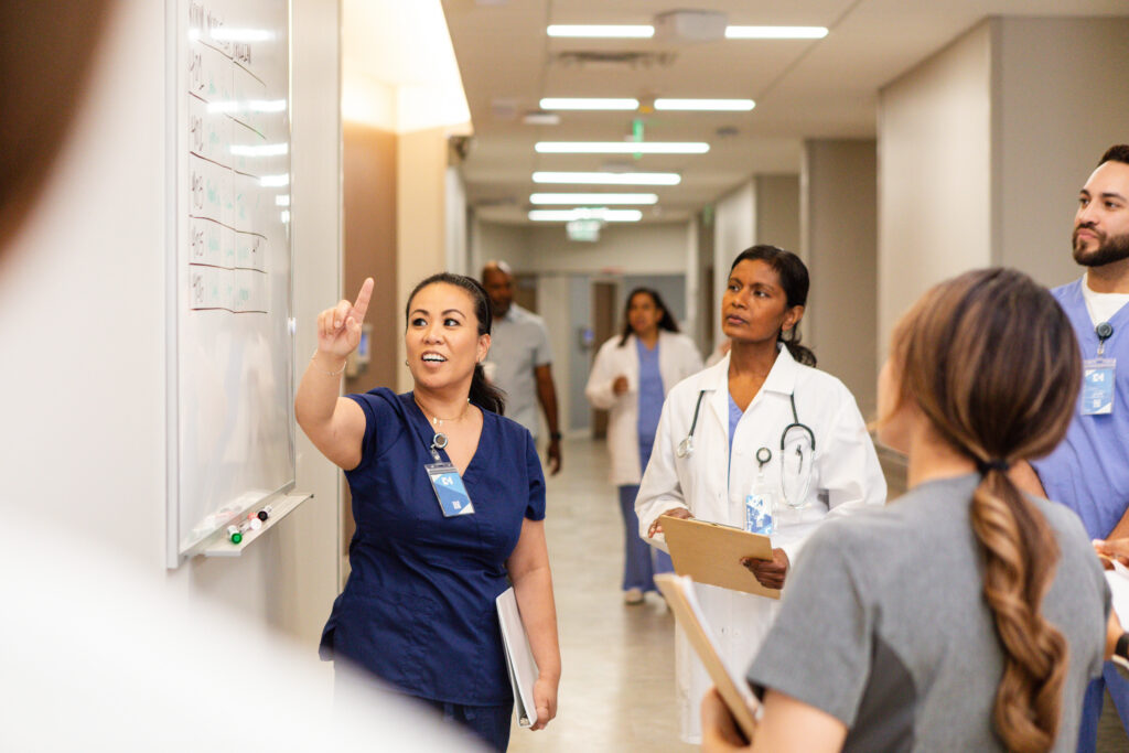 Diverse team of nurses and doctors collaborating during a clinical huddle at a hospital whiteboard, representing Guidehealth's Human Healthguides and nursing teams delivering coordinated whole-person care.
